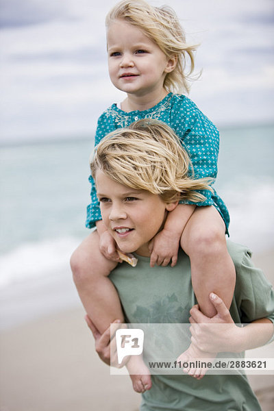 Junge  der seine Schwester auf den Schultern am Strand trägt.