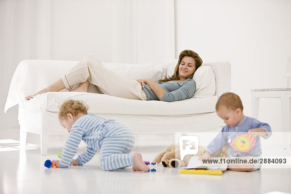 Babies playing with toys and their mother resting on a couch