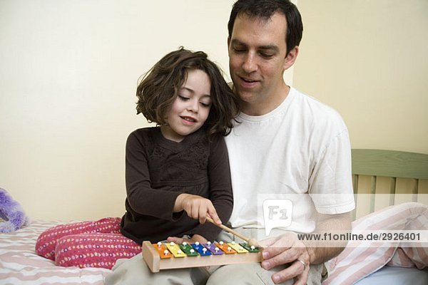 A young girl sitting with her father and playing the xylophone