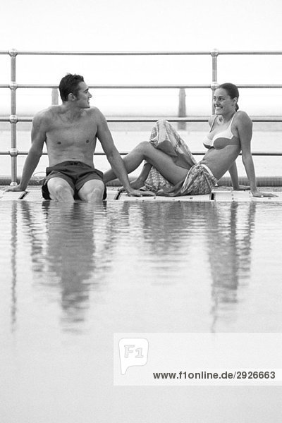Junges Paar sitzt zusammen am Pool und lächelt sich an.