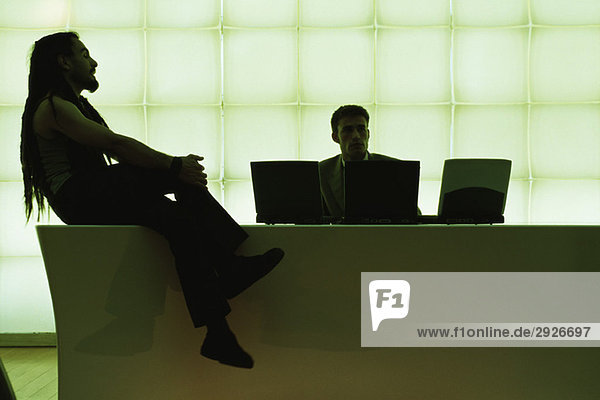 Mann mit Dreadlocks sitzt auf einem Schreibtisch mit gekreuzten Beinen und spricht mit einem Geschäftsmann über einen Laptop.