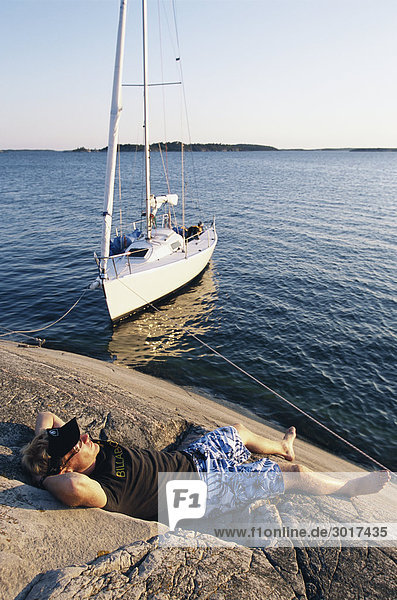 Ein Segelnboot und ein Mann auf einer Klippe Schweden Sonnenbaden.