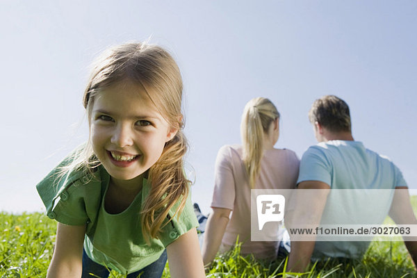  Parents sitting in meadow,  girl (6-7) smiling in foreground