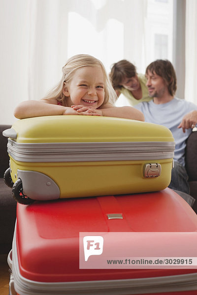 Mädchen (4-5) ruht Kinn auf Koffer,  Eltern im Hintergrund,  lächelnd,  Portrait