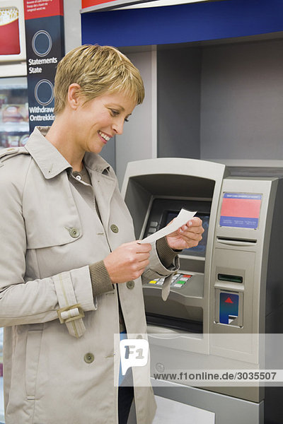 Frau liest einen Transaktionsschein bei einem Geldautomaten und lächelt