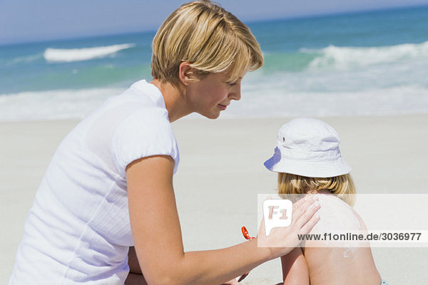 Frau  die Sonnencreme auf ihre Tochter am Strand aufträgt.