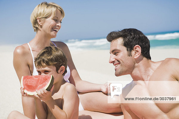 Junge isst eine Wassermelone mit seinen Eltern am Strand.