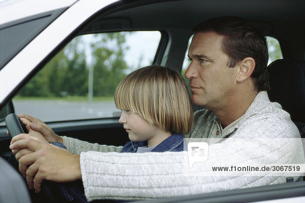 Mann fährt Auto mit jungem Sohn auf dem Schoß  beide halten das Lenkrad.