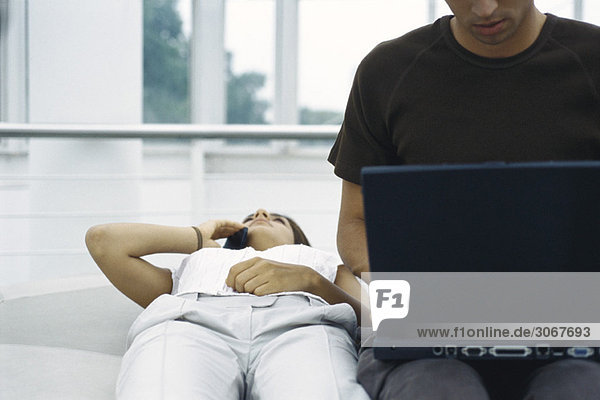 Junge Frau auf dem Rücken liegend mit dem Handy  Mann mit dem Laptop neben ihr