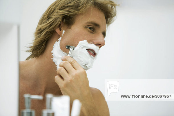 Mann rasiert sich mit Rasiermesser und schaut sich selbst im Spiegel an.