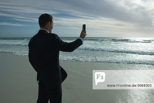 Geschäftsmann am Strand stehend  fotografiert Meer mit Fotophon