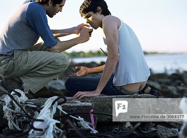 Young man lighting second young man's cigarette  on junky shore