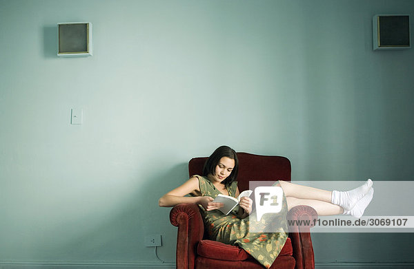 Frau sitzt im Sessel und liest Buch  trägt Kleid und Socken  volle Länge