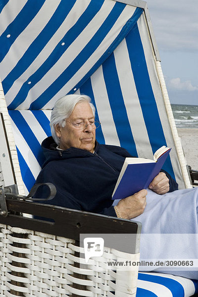 Ein älterer Mann sitzt in einem Strandstuhl mit Kapuze und liest.