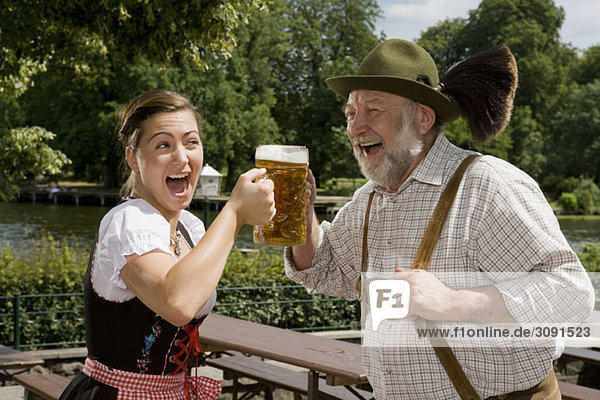 Ein traditionell gekleideter deutscher Mann und eine deutsche Frau in einem Biergarten mit Toastgläsern.