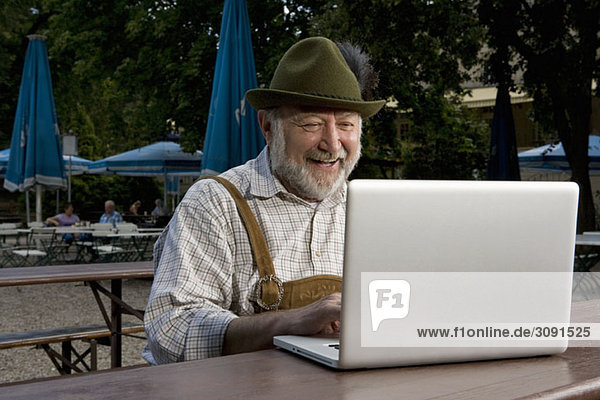 Ein traditionell gekleideter deutscher Mann in einem Biergarten mit einem Laptop