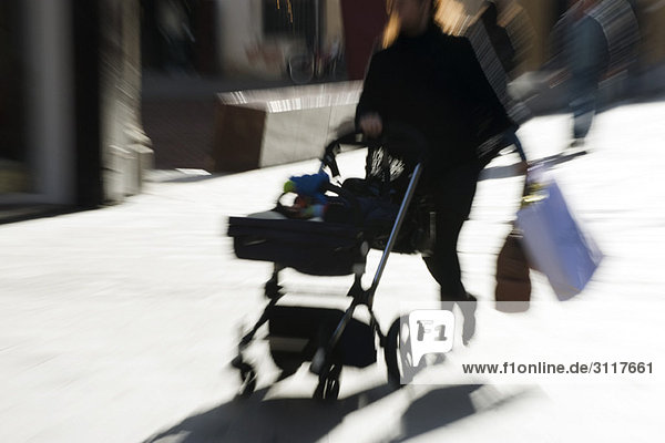 Pedestrian pushing baby carriage on sidewalk  carrying shopping bags  blurred