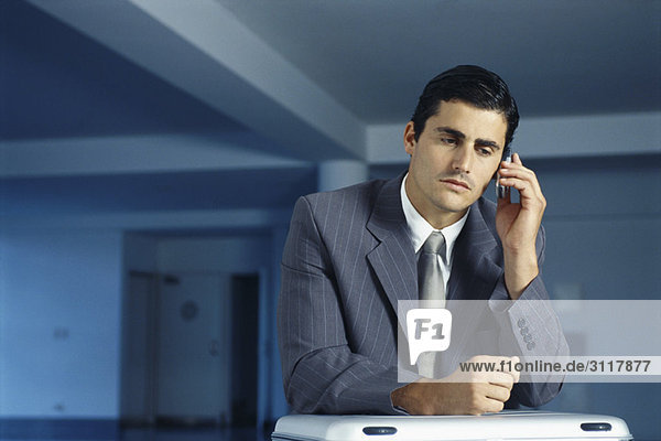 Businessman sitting with briefcase on lap in empty office using cell phone