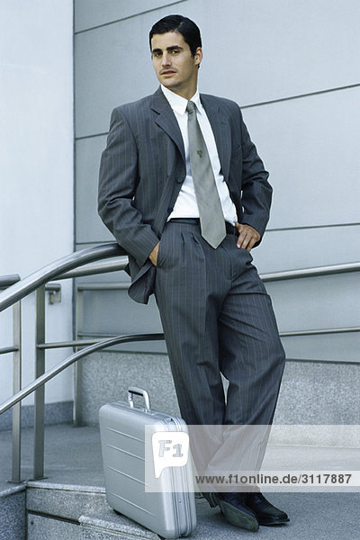 Businessman leaning against railing  briefcase on sidewalk at feet