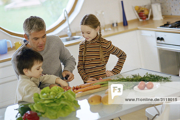 Vater mit Kindern in der Küche  hilft dem kleinen Sohn beim Gemüse schneiden