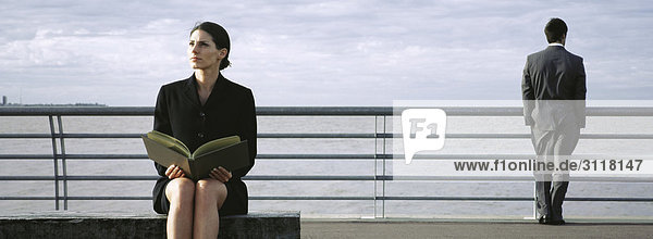 Geschäftsfrau auf Bank am Wasser mit offenem Buch auf dem Schoß  Mann am Geländer mit Blick in den Hintergrund