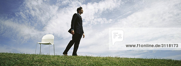 Businessman walking barefoot in grassy field,  empty chair nearby