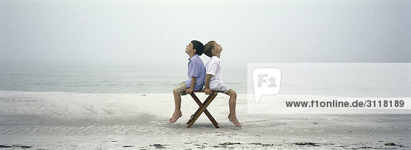 Zwei Jungen sitzen Rücken an Rücken auf einem Stuhl am Strand.