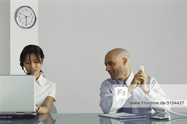 Büroangestellte  die am Schreibtisch sitzt  Telefon hält  den Hörer abdeckt und mit einer Mitarbeiterin spricht.