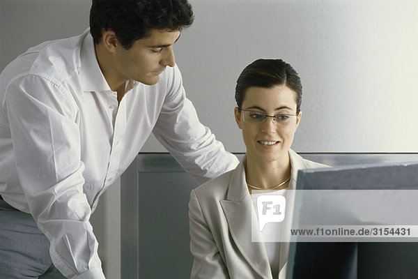 Junge Berufstätige lächelnd auf den Computerbildschirm blickend  während sich der Kollege über die Schulter lehnt.