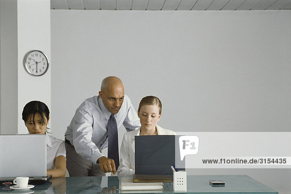 Professioneller Mann  der hinter einer jungen Kollegin steht und auf ihren Computerbildschirm zeigt.