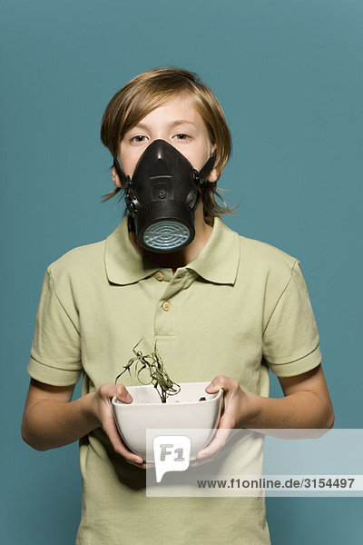 Junge mit Gasmaske  der eine verwelkte Topfpflanze hält.