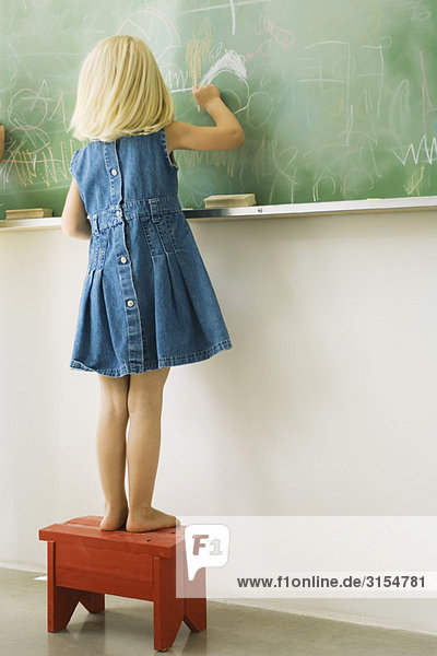 Kleines Mädchen auf Hocker stehend  auf Tafel kritzelnd  Rückansicht