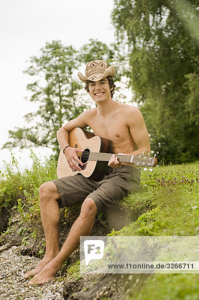 Starnberger See  Junger Mann sitzt am Seeufer und spielt Gitarre  lächelnd  Portrait