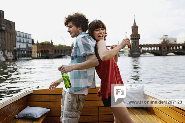 Germany  Berlin  Young couple on motorboat  holding bottles  standing back to back