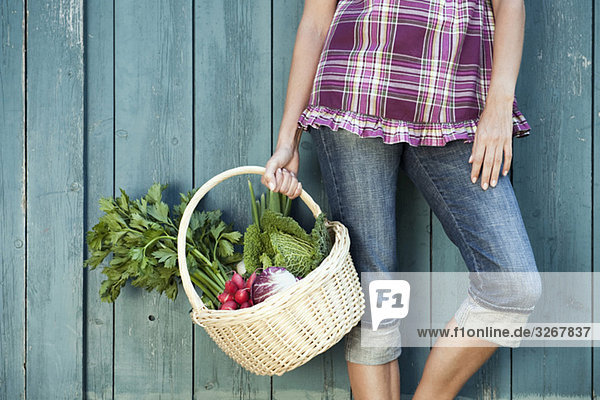 Frau lehnt sich an das Scheunentor und hält einen Korb mit frischem Gemüse.
