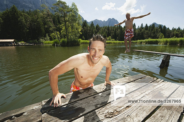 Italy  South Tyrol  Man in foreground leaning on jetty  senior man in background jumping into lake  portrait