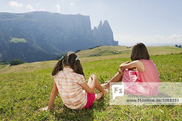 Italy  South Tyrol  Seiseralm  Two girls (6-7) (10-11) sitting in meadow  rear view