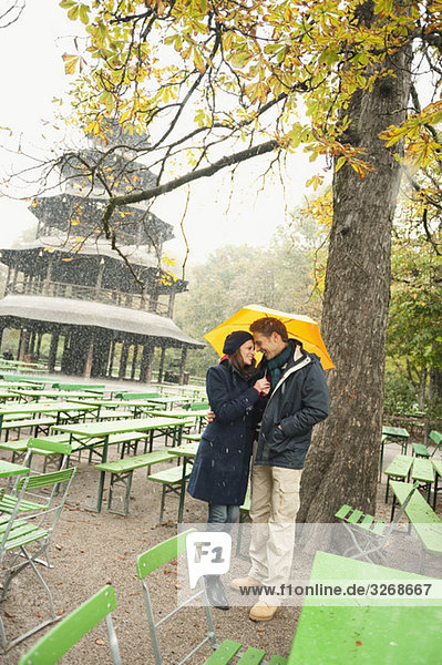 Deutschland  Bayern  München  Englischer Garten  Paar im verregneten Biergarten mit Regenschirm  Chinesischer Turm im Hintergrund  Portrait