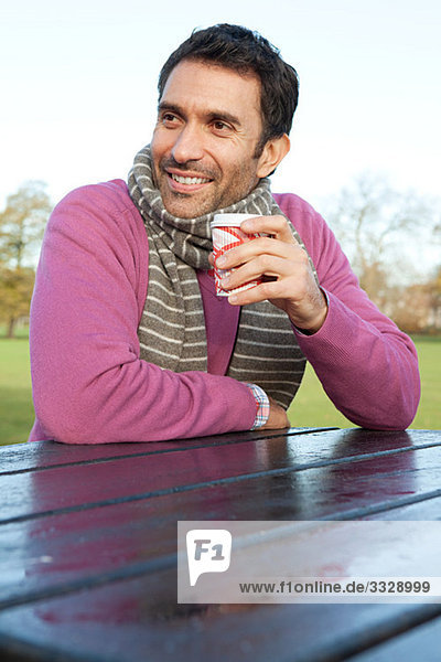 Ein Mann  der an einem Parktisch sitzt und ein heißes Getränk hält.