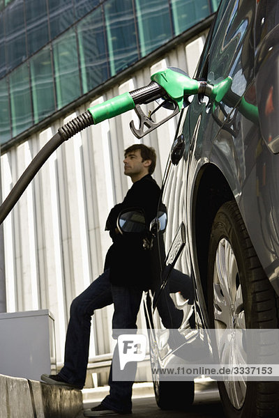 Mann an der Tankstelle lehnt sich an das Auto und wartet darauf  den Tankvorgang zu beenden.