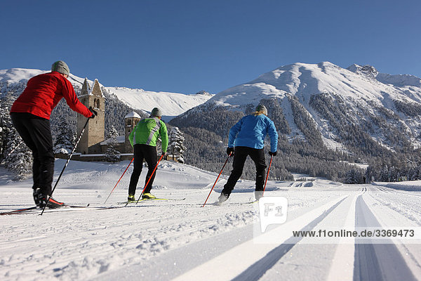 10870407  Winter sports  Switzerland  cross-country skiing  group  three  canton Graubunden  Grisons  Bundnerland  cross-country  skiing  Engadin  Oberengadin  persons  people  blue  red  green  church San Gian  cross-country skiing cross-country trail  tracks  traces  back view