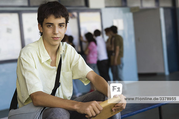 Männlicher Gymnasiast sitzt auf einer Bank und hält ein gebundenes Buch.