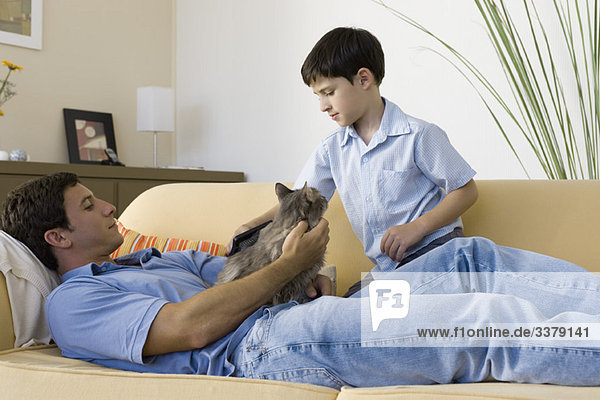Mann entspannt mit Katze auf dem Schoß  junger Sohn bürstet Katze