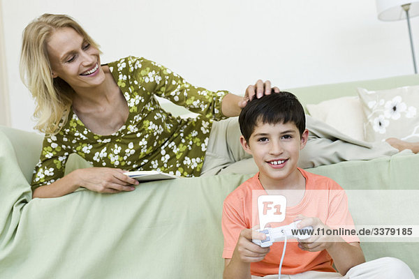 Young boy sitting on floor playing video game  mother relaxing on sofa