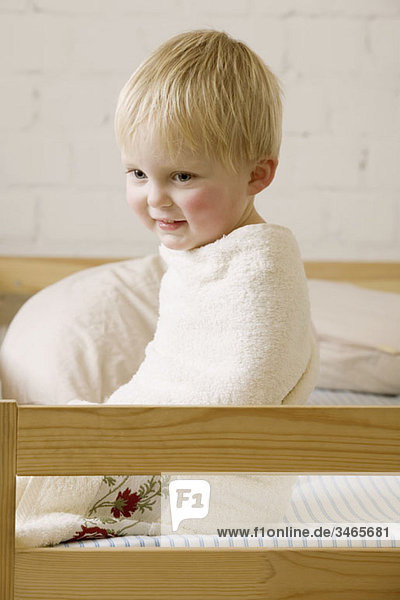 Ein fröhlicher kleiner Junge in ein Handtuch gewickelt  das auf einem Bett sitzt.