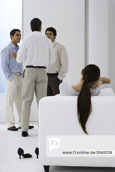 Frau entspannt im Sessel  während männliche Kollegen im Hintergrund plaudern