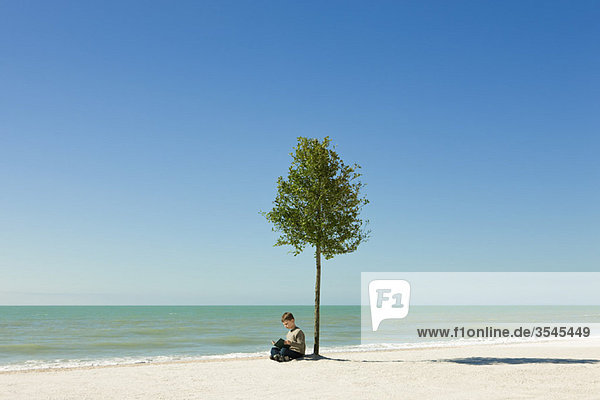 Junge liest Buch unter einem Baum am Strand