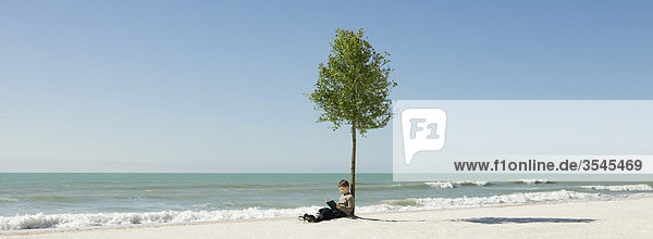 Junge liest Buch unter einem Baum am Strand