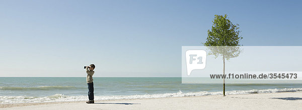 Junge steht am Strand und schaut mit einem Fernglas auf das Meer.