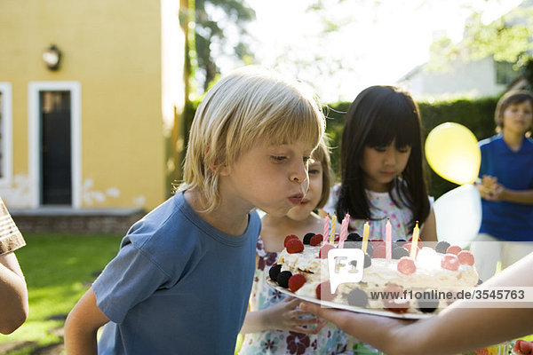 Junge bläst Kerzen auf Geburtstagskuchen bei einer Geburtstagsparty im Freien aus.